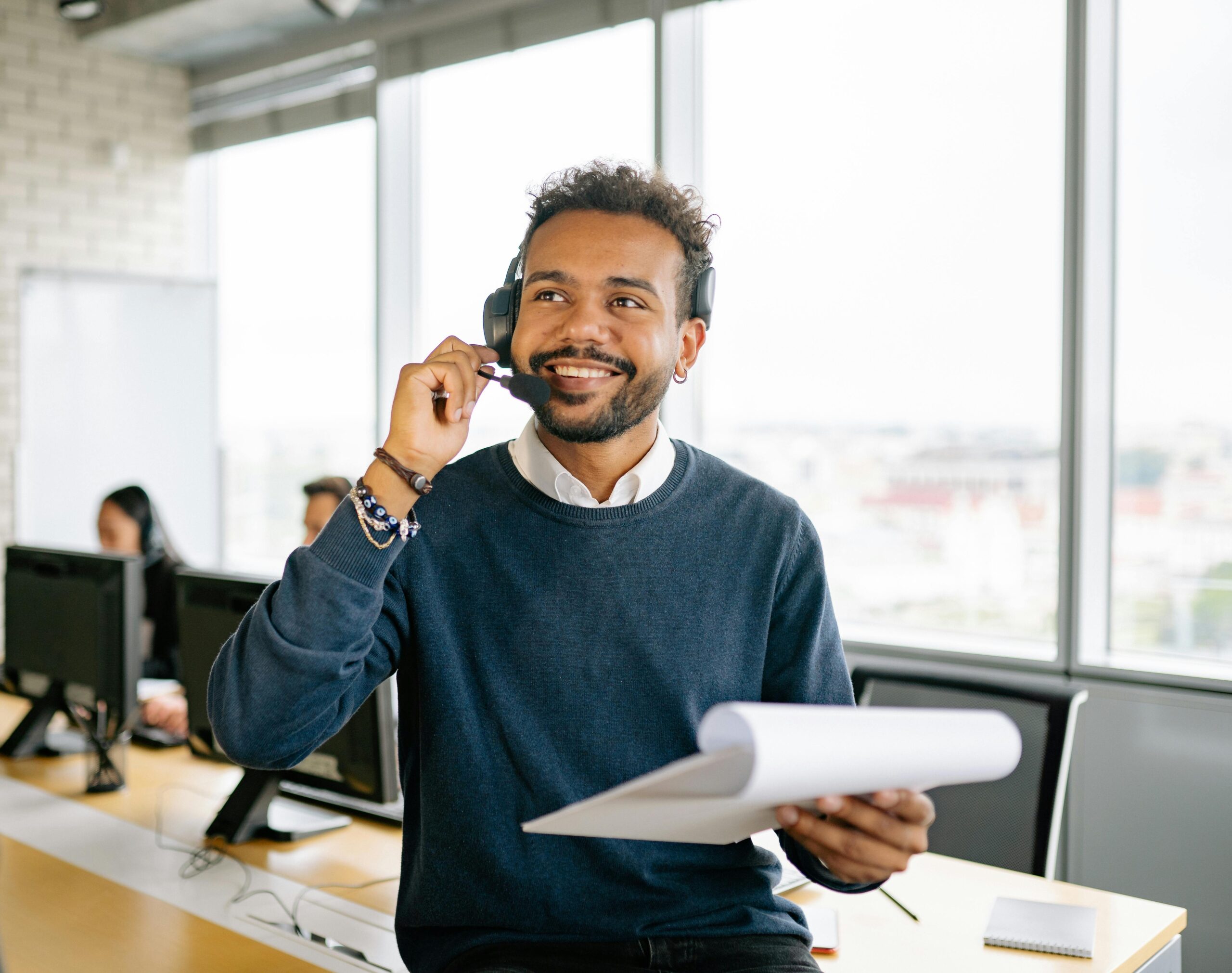 A cheerful man wearing a headset and holding a clipboard at a modern office workspace.